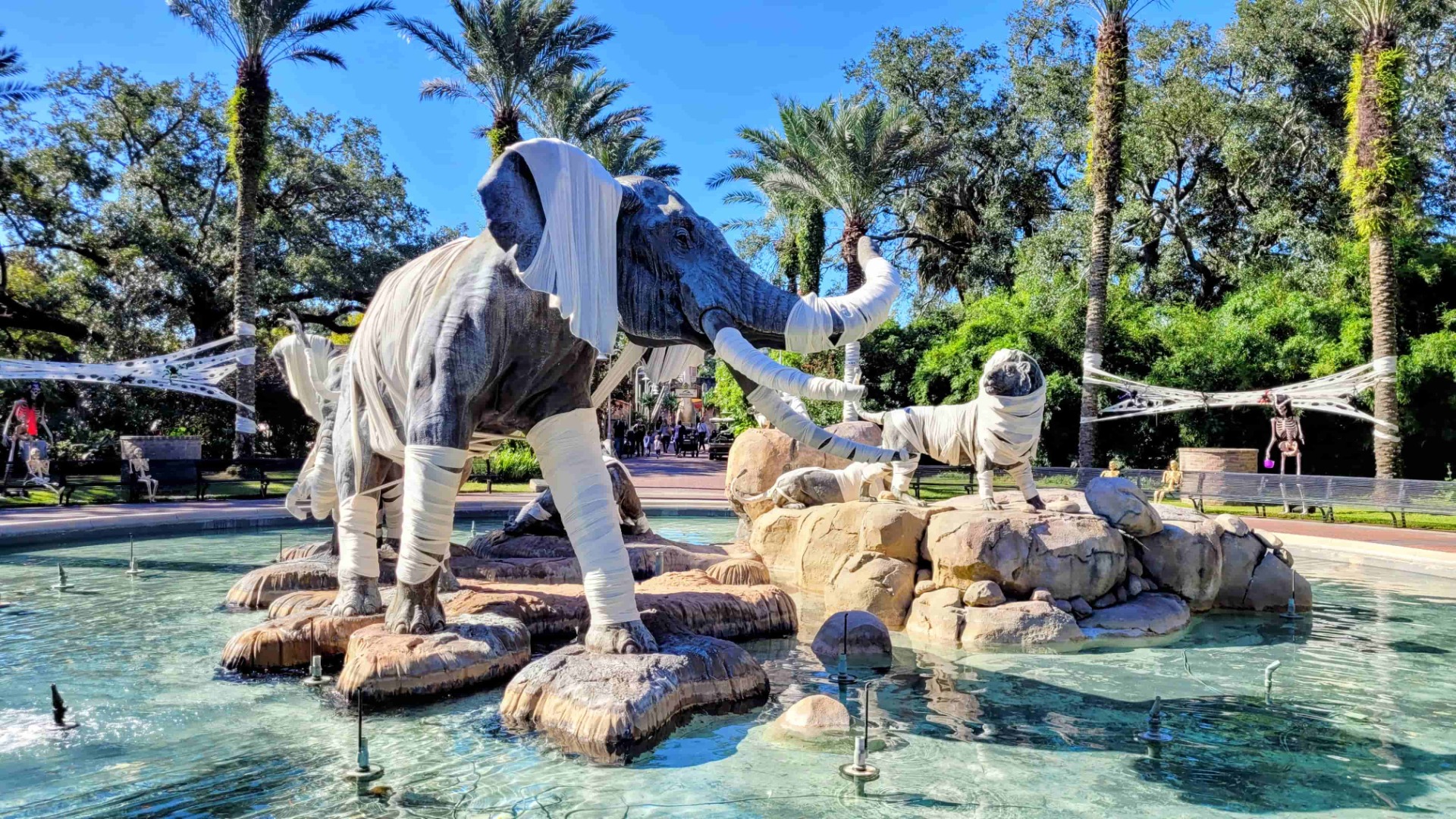 Statuen in weißen Gewändern stehen im Wasserbrunnen, umgeben von Palmen und Sonnenschein im Park.