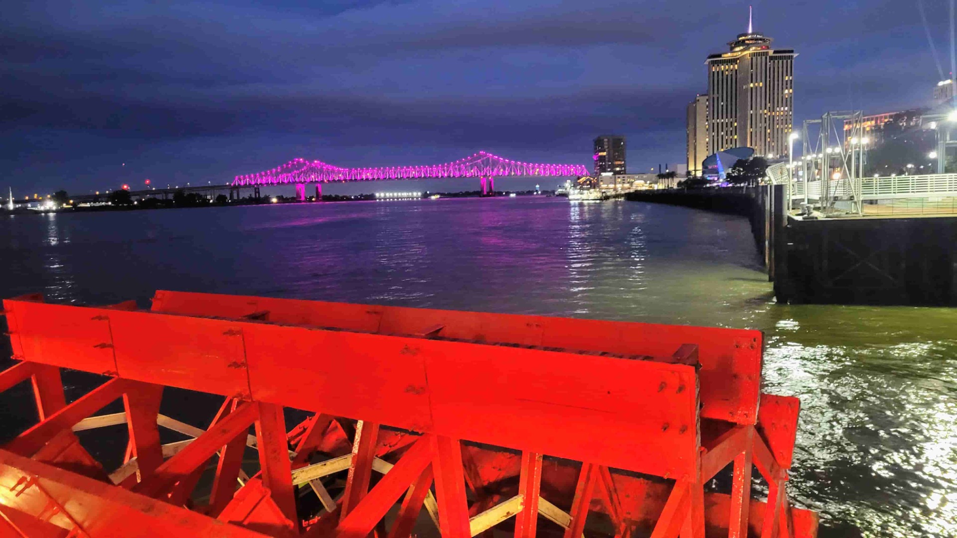 Roter Geländerblick auf den Fluss bei Nacht, beleuchtete Brücke und Hochhaus mit Lichtern im Hintergrund.