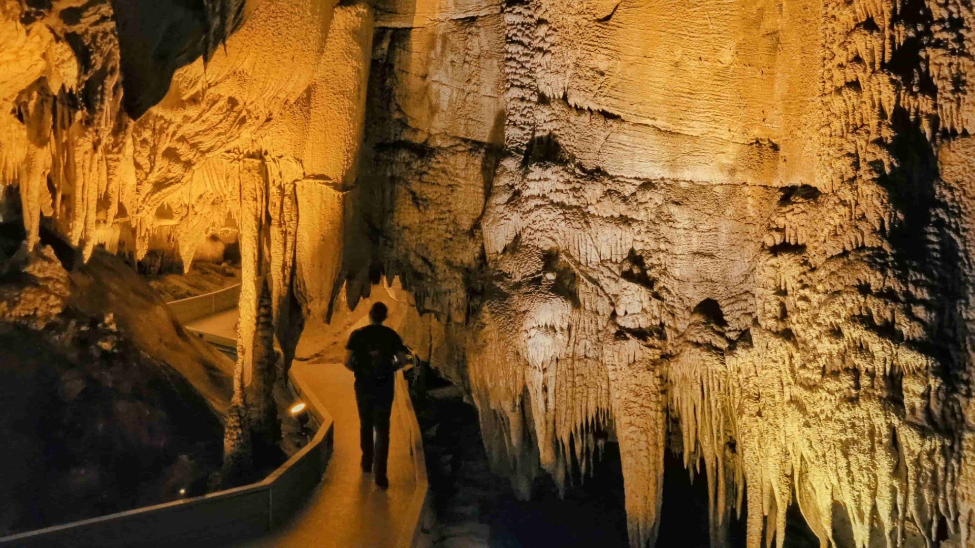 Person geht durch eine Tropfsteinhöhle, goldene Wände mit Stalaktiten und beleuchtetem Weg.