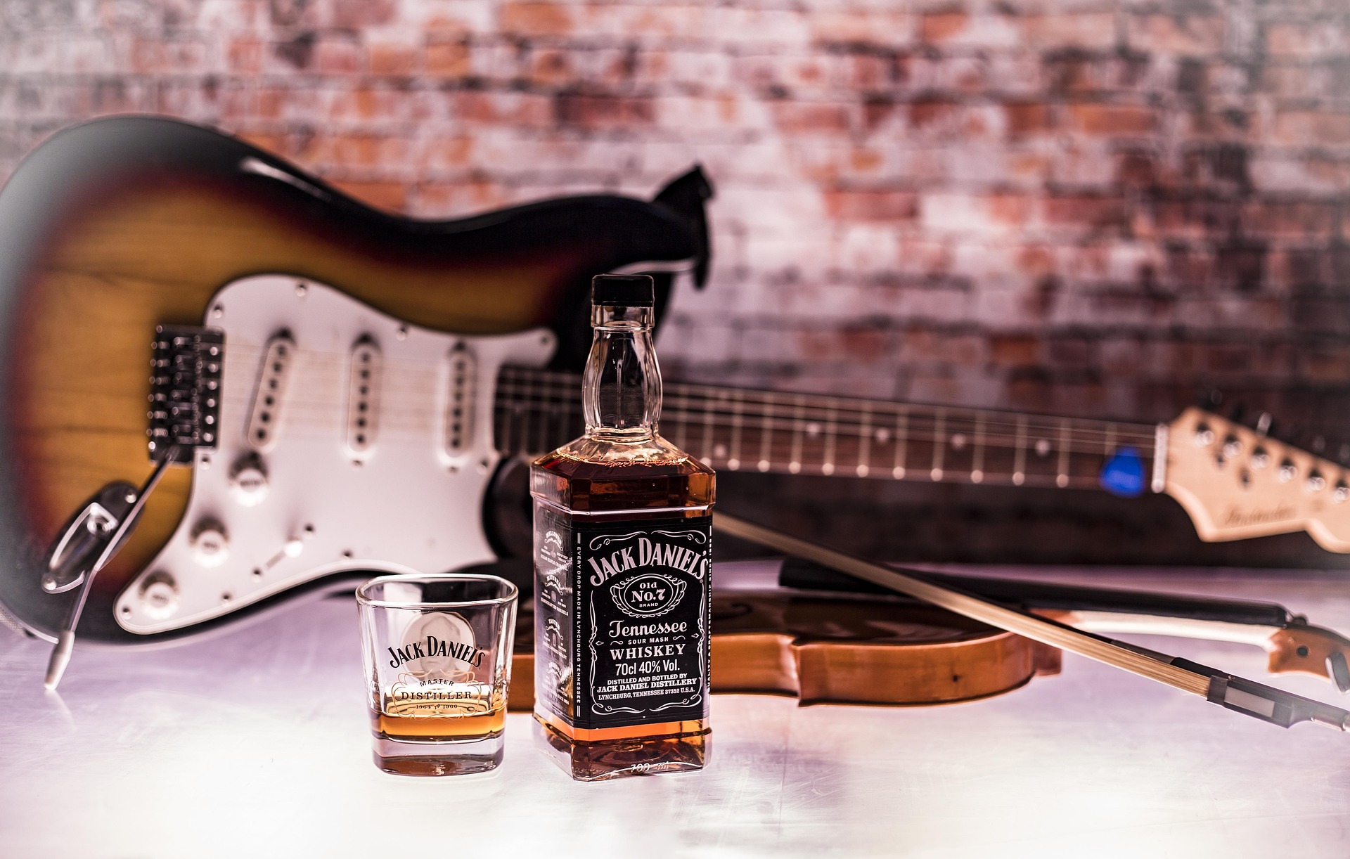 Jack Daniel’s Flasche und Glas mit Whiskey vor Gitarren im Hintergrund, Ziegelwand und Saiten sichtbar.